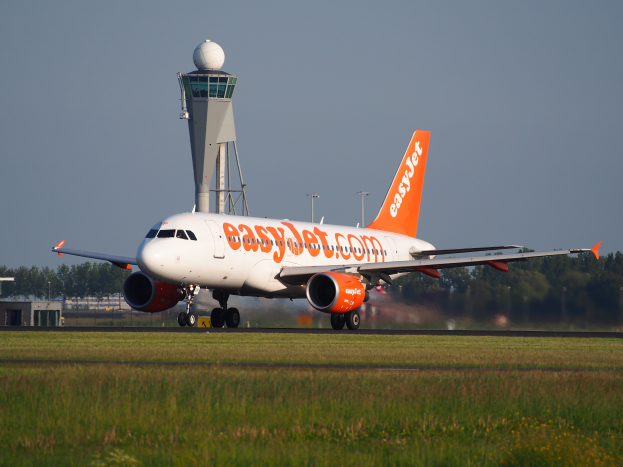 Ein EasyJet Airbus A320-200 Flugzeuge auf dem Rollfeld am Frankfurt Airport, mit Gras, einer Kontrollturm, B├Ąumen, Geb├Ąuden und einem klaren blauen Himmel im Hintergrund.