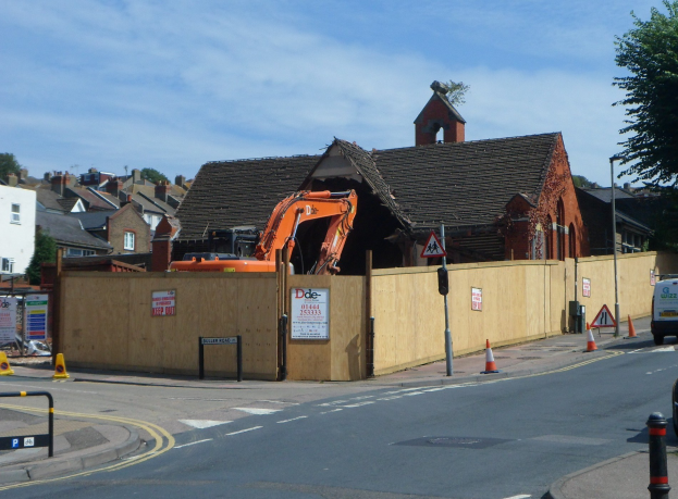 Straße mit Fahrzeugen, Verkehrsleitkegeln, Pfosten, Schildern und einem Zaun, die zu Gebäuden im Hintergrund mit Bäumen, Wolken und Himmel führt; ein Bagger und ein abbruchreifes Gebäude am Straßenrand.