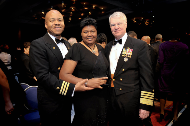 Zwei Männer in schwarzen Blazern und weißen Hemden stehen neben einer Frau in einem schwarzen Kleid, die eine Auszeichnung hält, auf einem roten Teppich, während andere im Hintergrund sitzen und stehen unter beleuchteter Decke.