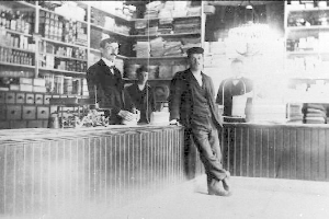 Ein Schwarz-Weiß-Foto von drei Männern, die an einem Tresen in einem Laden stehen, mit verschiedenen Gegenständen auf dem Tresen und einem Regal mit Büchern und anderen Gegenständen im Hintergrund während eines Raubüberfalls.