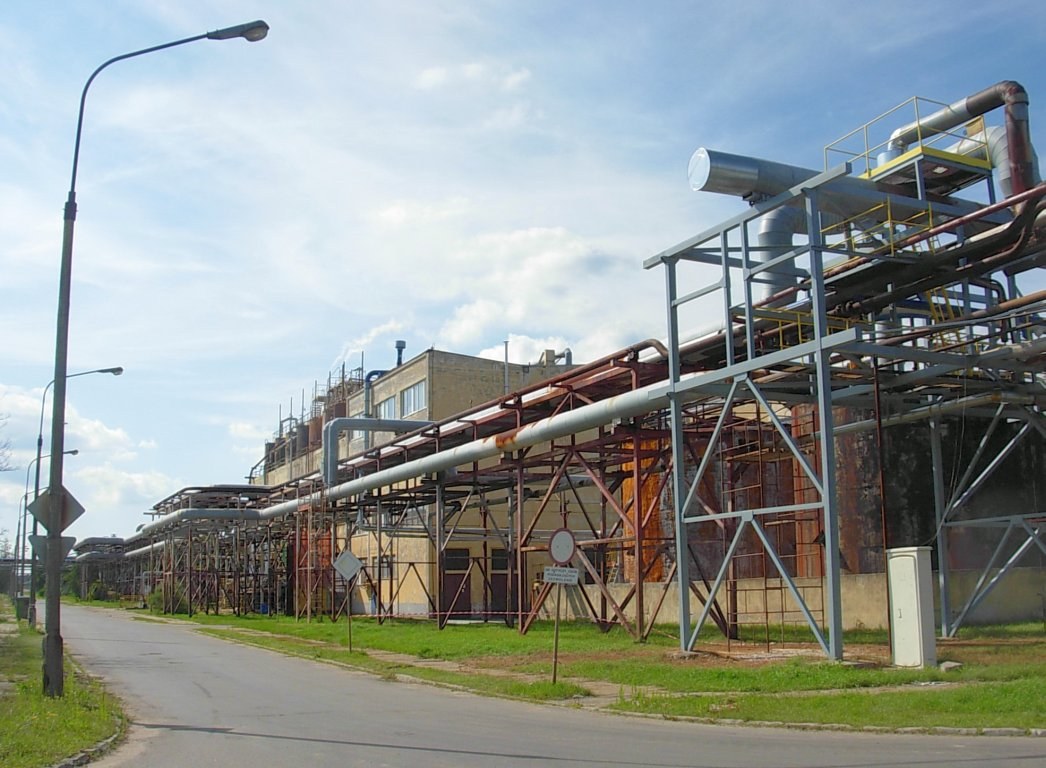 Große Fabrik mit ausgedehnten Rohrleitungen, umgeben von städtischer Infrastruktur und Grünflächen unter einem bewölkten Himmel.