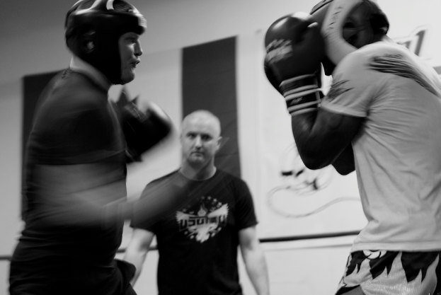 Zwei Boxer im Ring mit Helmen und Handschuhen kämpfen gegeneinander, während ein Schiedsrichter zwischen ihnen steht und ein Banner im Hintergrund zu sehen ist.