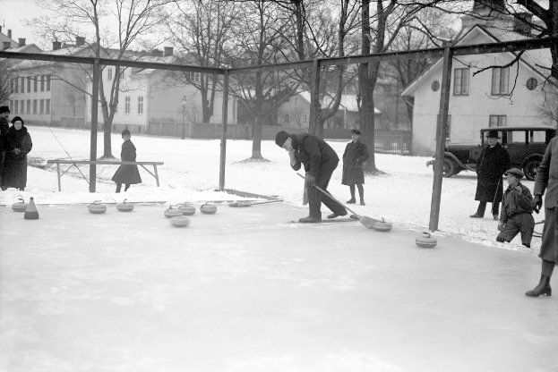 Schwarzes Bild von Menschen, die Curling auf einer Eisbahn spielen, umgeben von einem Zaun, einer Bank, Bäumen, Gebäuden und einem geparkten Fahrzeug im Hintergrund.