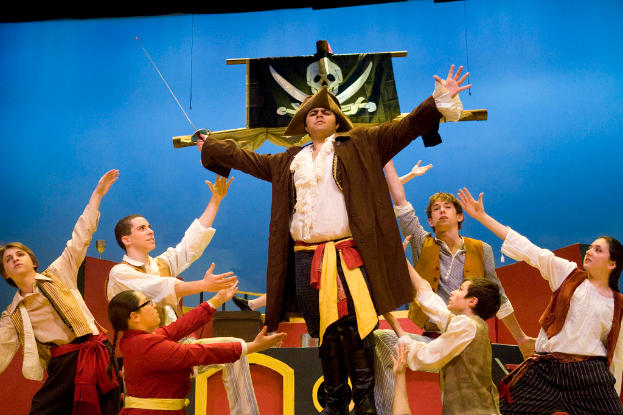 Ein Mann hält ein Nadelsschwert in den Händen, umgeben von Menschen mit erhobenen Händen, mit einem blauen Hintergrund und einer Fahne im Hintergrund.