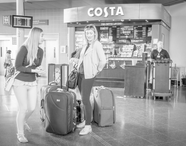 Zwei Frauen mit Gepäcktaschen vor einem Kaffeehaus an einem Ort, der wie ein Flughafen aussieht.