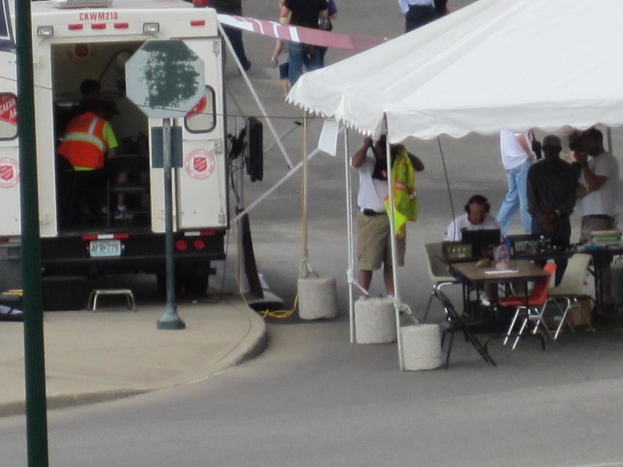 Ein Krankenwagen steht auf der linken Seite einer Stra├če, w├Ąhrend auf der rechten Seite ein Zelt viele Menschen sch├╝tzt, mit einem Tisch und einem Stuhl in der N├Ąhe.