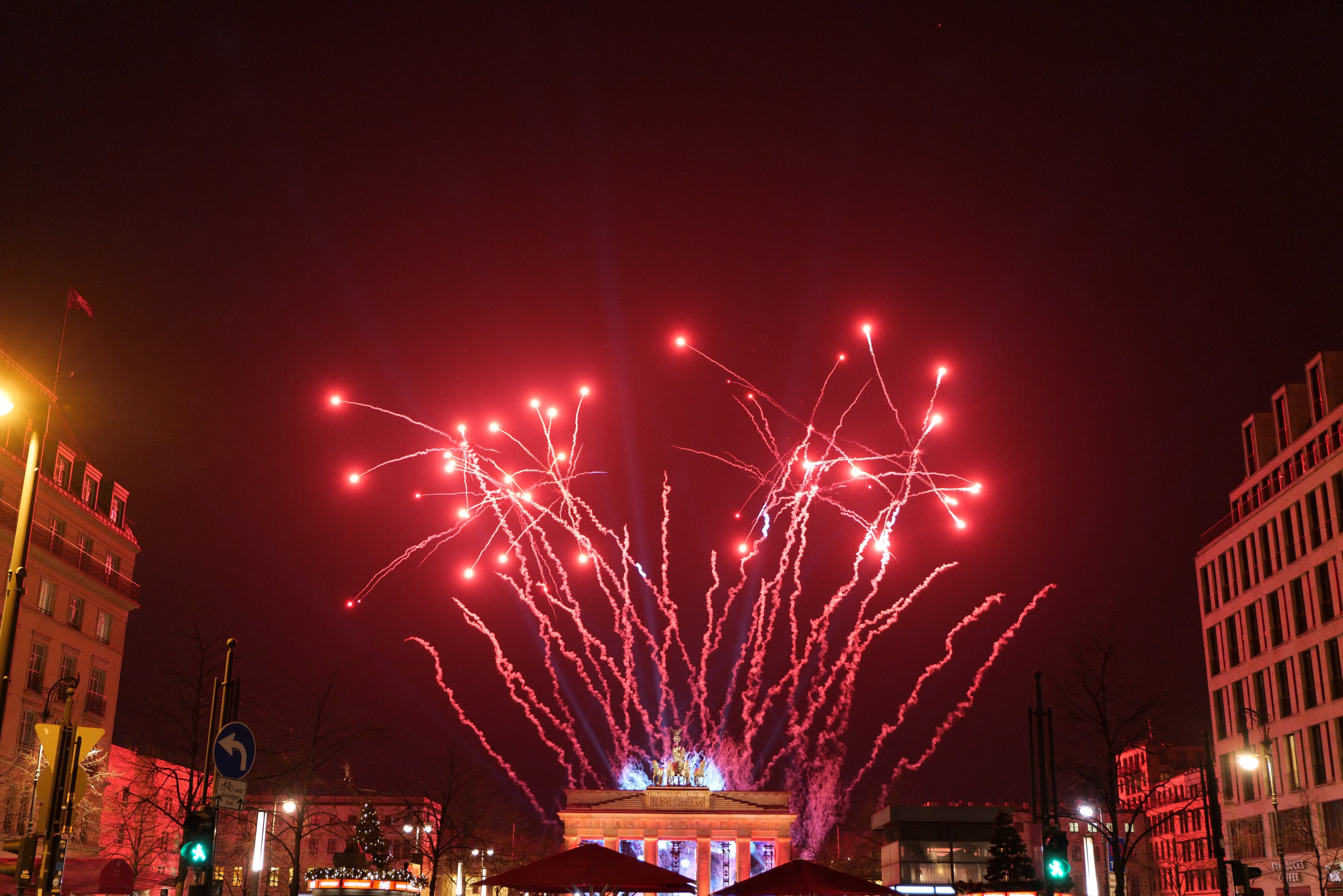 Eine belebte Stadtstraße bei Nacht während des Neujahrsfeier in Berlin, mit Gebäuden, Bäumen, Laternen, Ampeln, Schildern, Zelten, Menschen und einem prächtigen Feuerwerk am Himmel.