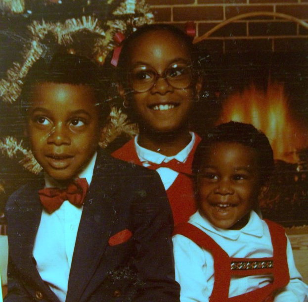 Drei Kinder lachen vor einem Weihnachtsbaum mit einem Kamin aus Holz auf der rechten Seite.