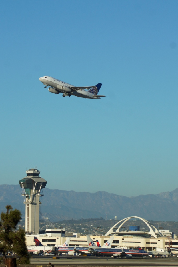 Eine Flughafenszene mit einem weißen Kontrollturm im Vordergrund, mehreren geparkten Flugzeugen, entfernten Gebäuden und einem Flugzeug am Himmel.