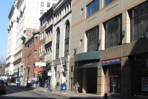 Eine belebte Stadtstraße mit hohen Gebäuden, fahrenden Fahrzeugen, Fußgängern auf dem Gehweg, einem Feuerhydranten im Vordergrund, Straßenlaternen, Bäumen im Hintergrund und einem klaren blauen Himmel.