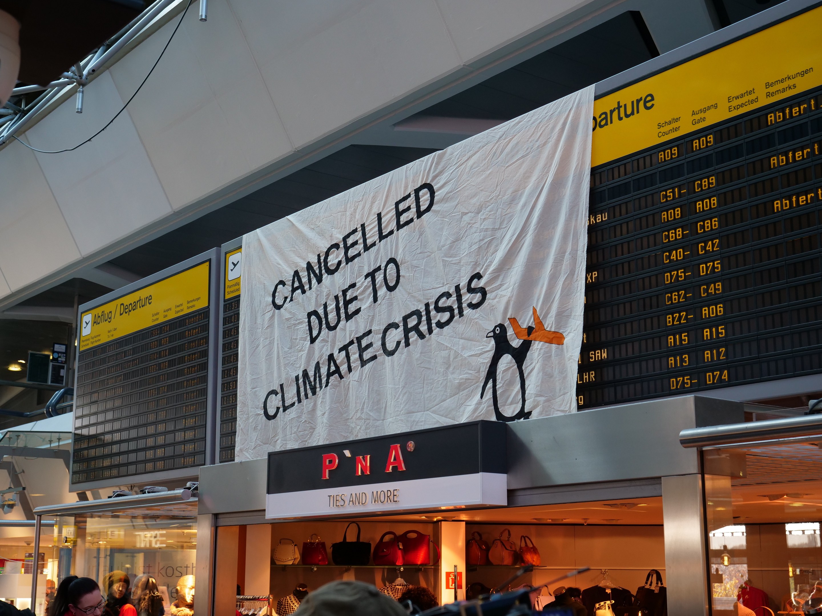 Gruppe von Menschen vor einer Abflugtafel mit einem Banner "Aufgrund der Klimakrise gestrichen"