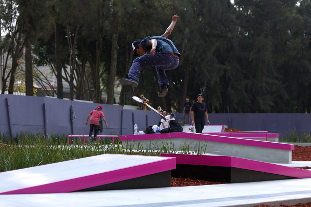 Ein Skateboarder, der einen Sprung auf einer Rampe ausführt, während eine Gruppe von Zuschauern zuschaut, einige halten Skateboards, mit verstreuten Taschen, Flaschen und anderen Gegenständen in der Nähe, vor einem Hintergrund aus Bäumen, Gebäuden und einem klaren blauen Himmel.