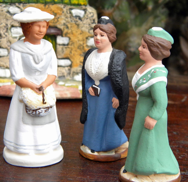 Drei Frauen-Skulpturpuppen auf einer Holzplanke mit Spielzeughäusern im Hintergrund.