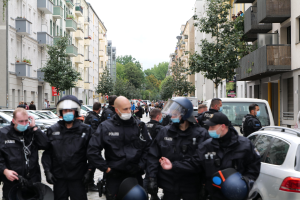 Eine Gruppe von Polizisten in Masken und Helmen, die eine von parkenden Autos gesäumte Straße entlanggehen, flankiert von Gebäuden mit Balkonen und Geländern, mit Bäumen und einem klaren Himmel im Hintergrund.