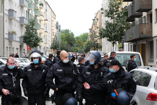 Eine Gruppe von Polizisten in Masken und Helmen, die eine von parkenden Autos gesäumte Straße entlanggehen, flankiert von Gebäuden mit Balkonen und Geländern, mit Bäumen und einem klaren Himmel im Hintergrund.
