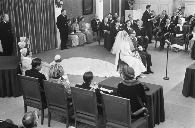 Schwarzes und weißes Foto einer Hochzeitszeremonie mit sitzenden Gästen, einem Mann, der in ein Mikrofon spricht, einer Frau mit einem Blumenstrauß, einem Podium mit einem Mikrofon, Blumengestecken, einem Vorhang, einem gerahmten Foto an der Wand und einer Tür im Hintergrund.