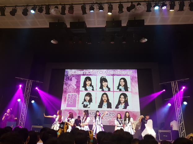 Eine Gruppe von Menschen, die auf einer hell erleuchteten Bühne performen, mit einer Menge darunter, die einen Sänger mit Mikrofon und einen Musiker mit Instrument zeigt, während ein großer Bildschirm im Hintergrund Bilder der K-Pop-Gruppe BK48 anzeigt.