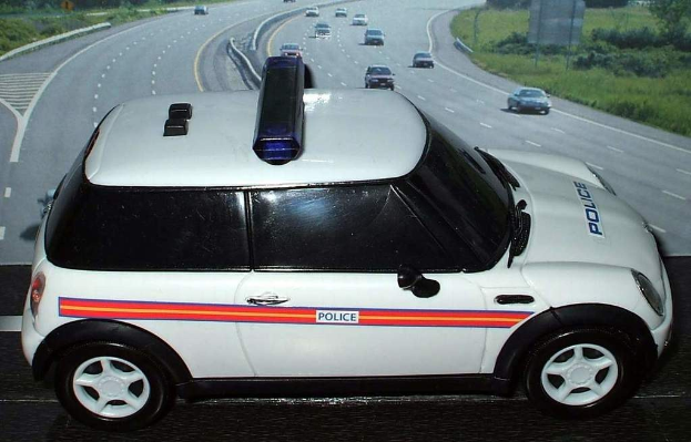 Weißer Polizeiwagen mit "Polizei" Aufschrift vor einer Reihe fahrender Fahrzeuge auf einer Straße mit Gras und Bäumen außerhalb einer Stadt.