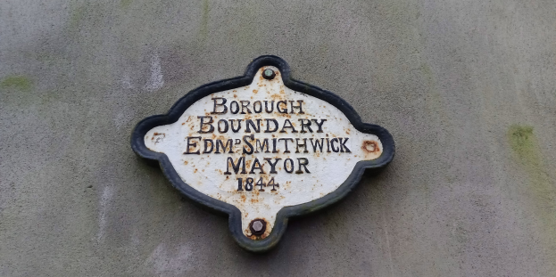 Metallplatte an der Außenseite eines Gebäudes mit schwarzem Hintergrund und weißer fettgedruckter Schrift, die "Stadtteilgrenze Edm Smithwick Bürgermeister" lautet.
