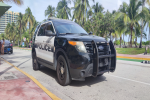 Polizeiauto am Straßenrand mit Menschen, Fahrrädern, Gras, Bäumen, Gebäuden und einem klaren blauen Himmel im Hintergrund; das Vorderrad des Autos ist sichtbar.
