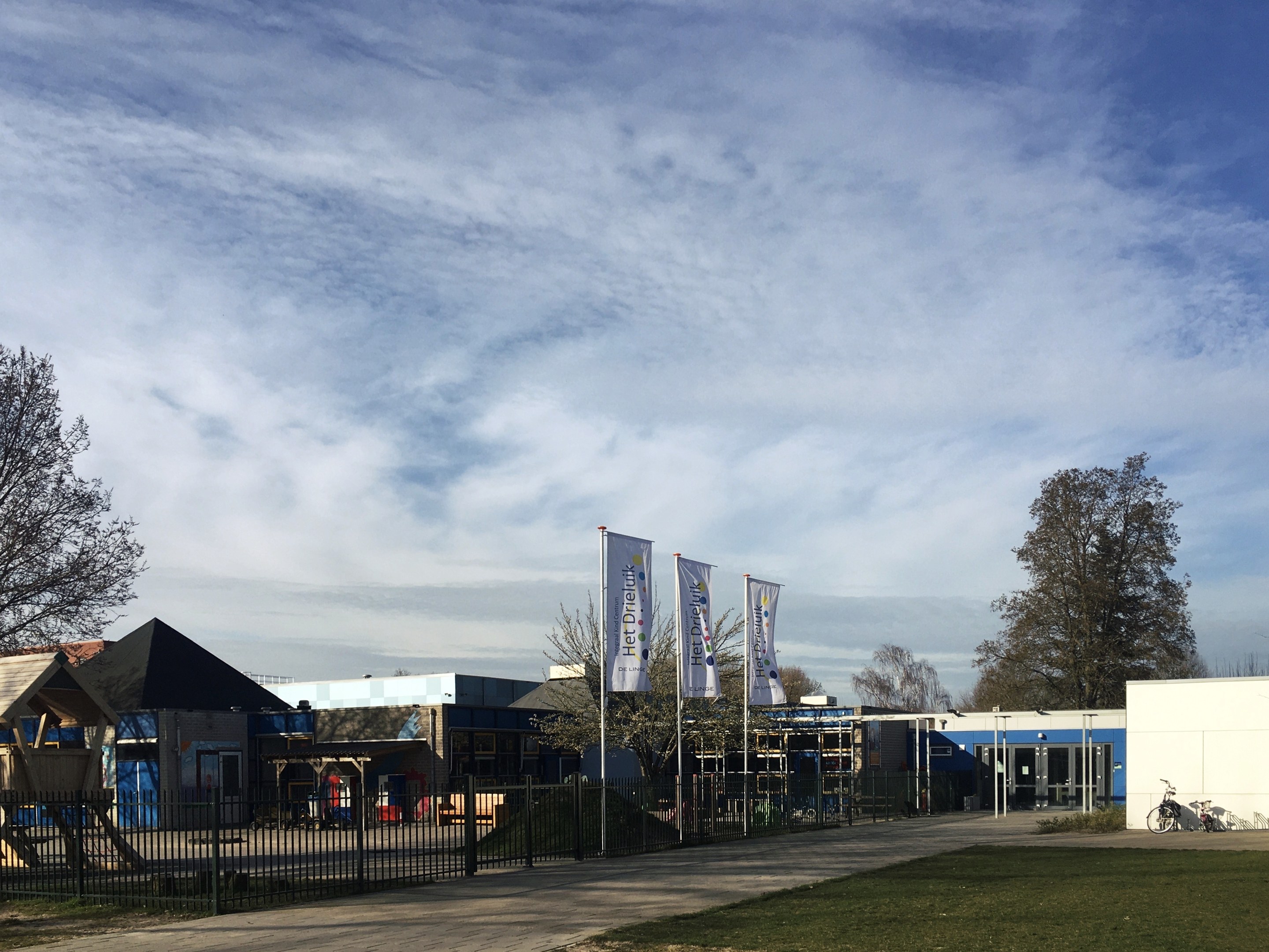 Großes Gebäude mit einem grasbewachsenen Bereich davor, umgeben von einem Metallzaun, Bäumen, Bannern, einem Fahrrad, nahegelegenen Häusern und einem bewölkten Himmel, identifiziert als neuer Spielplatz an einer Schule.
