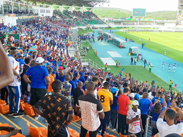 Eine große Menschenmenge versammelt sich vor einem eingezäunten Fußballfeld, mit einigen auf Stühlen sitzend und anderen stehend, unter einem klaren blauen Himmel mit Hügeln im Hintergrund, in Erwartung des 2019 African Cup of Nations-Spiels.
