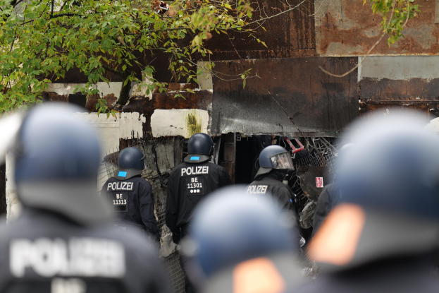 Eine Gruppe von Polizisten in schwarzen Uniformen und Helmen, die vor einem mit Maschendrahtzaun umgebenen Gebäude stehen, mit Bäumen im Hintergrund.