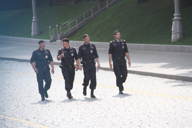 Drei Polizisten in Uniform und mit Mützen gehen eine Straße entlang mit Pfählen, Geländern, Treppen und Gras im Hintergrund.