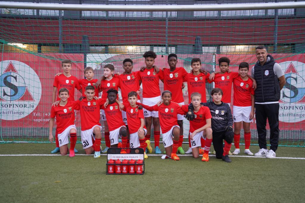 Eine Gruppe junger Männer in roten T-Shirts und schwarzen Shorts steht auf einem Fußballfeld vor einer Kiste roter Gegenstände, im Hintergrund sind Banner mit "FC Rot Schwarz Eglaute - Champions League" zu sehen.