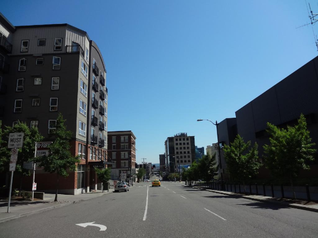 Eine Stadtansicht mit Fahrzeugen auf einer Straße, Bäumen, Pfählen auf dem Gehweg, Gebäuden, mit dem Himmel oben und der Straße unten.