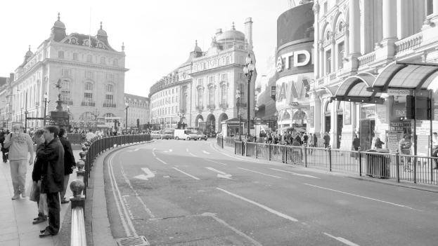 Eine Straße mit weißen Linien, Pfeilschildern, einem Geländer auf der rechten Seite und Stahlabsperrungen auf der linken Seite, mit Menschen auf beiden Seiten, Straßenlaternen und Gebäuden im Hintergrund.