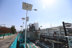 Großes industrielles Gebäude mit ausgedehnten Rohrleitungen und Solarmodulen auf dem Dach, umgeben von Sicherheitsgeländern, Strommasten, Straßenlaternen, Beschilderungen, Bäumen und vorbeifahrenden Fahrzeugen, vor einem Hügelpanorama unter einem klaren blauen Himmel, Teil der neuen Wasseraufbereitungsanlage Kaliforniens.