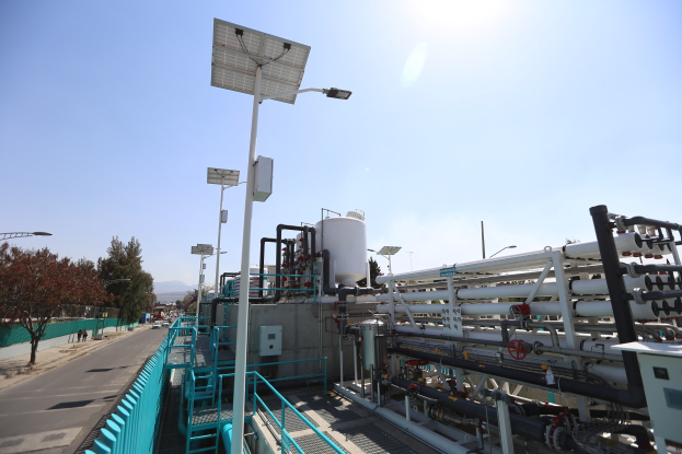 Großes industrielles Gebäude mit ausgedehnten Rohrleitungen und Solarmodulen auf dem Dach, umgeben von Sicherheitsgeländern, Strommasten, Straßenlaternen, Beschilderungen, Bäumen und vorbeifahrenden Fahrzeugen, vor einem Hügelpanorama unter einem klaren blauen Himmel, Teil der neuen Wasseraufbereitungsanlage Kaliforniens.