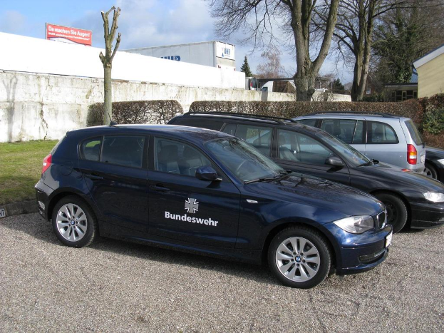 Mehrere Autos auf einem Weg umgeben von grünem Gras geparkt, mit Büschen, Bäumen und Gebäuden im Hintergrund, ein Schild mit Text und ein Logo sichtbar.
