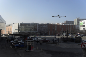 Eine belebte Stadtstraße mit parkenden Autos in einer Parkanlage neben hohen Gebäuden, beleuchtet von Straßenlaternen, mit ein paar Fußgängern und einem sichtbaren Himmel im Hintergrund.