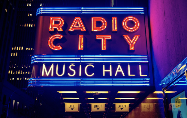 Außenansicht des Radio City Music Hall in New York City bei Nacht, hell erleuchtet und umgeben von hohen Gebäuden, mit einer "Radio City"-Tafel und zusätzlichen Tafeln auf der rechten Seite.