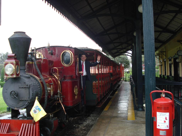 Ein alter roter Zug mit einem Mann in einem schwarzen Anzug, der an der Tür steht und lächelt, mit einer Stationshütte auf der rechten Seite und einem roten Feuerlöscher im Vordergrund.