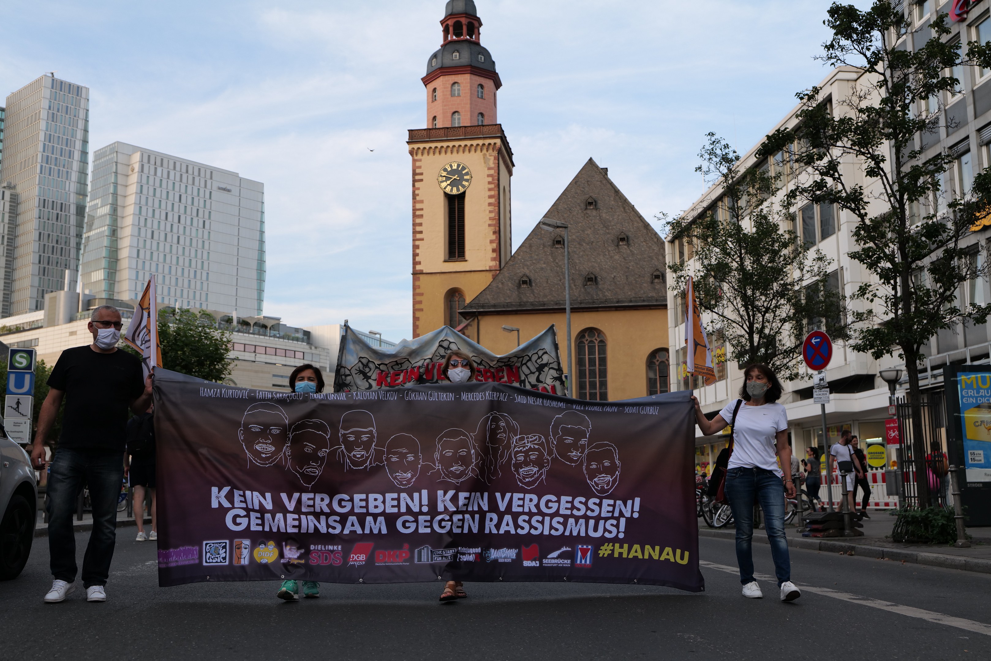 Eine Gruppe von Menschen in Masken, die eine Banner mit Text und Bildern tragen, geht eine Straße entlang, mit einem geparkten Auto auf der linken Seite, Gebäuden, Bäumen, Schildern, Masten, einem Uhrenturm und einem klaren blauen Himmel im Hintergrund.