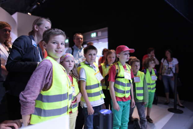 Eine Gruppe von Kindern in Sicherheitswesten und Namensschildern, mit einem Jungen, der links lächelt, auf einem Boden mit einem Pfahl rechts und einigen Leuten im Hintergrund.