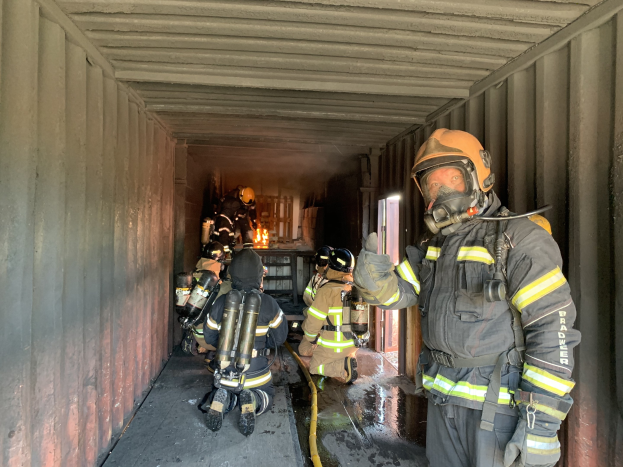 Feuerwehrleute in Schutzausrüstung, einschließlich Helmen, Handschuhen und Sauerstoffflaschen, innerhalb eines Seecontainers mit einem Feuerlöscher und anderer Ausrüstung im Hintergrund.