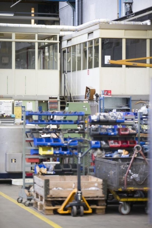 Ein großer Lagerraum voller verschiedener Ausrüstungsgegenstände wie Wagen, Schränke und eine Kiste, mit einer Wand aus Glasfenstern und Rohren im Hintergrund.