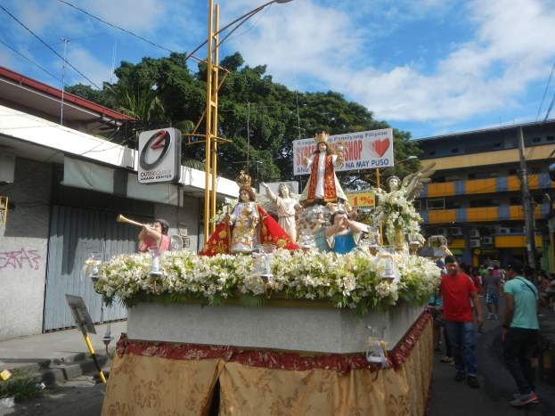 Ein Umzugswagen mit Statuen von Menschen, Blumen und anderen Gegenständen, begleitet von Texttafeln, Laternenmasten, Drähten, Gebäuden, Bäumen und einem bewölkten Himmel im Hintergrund.